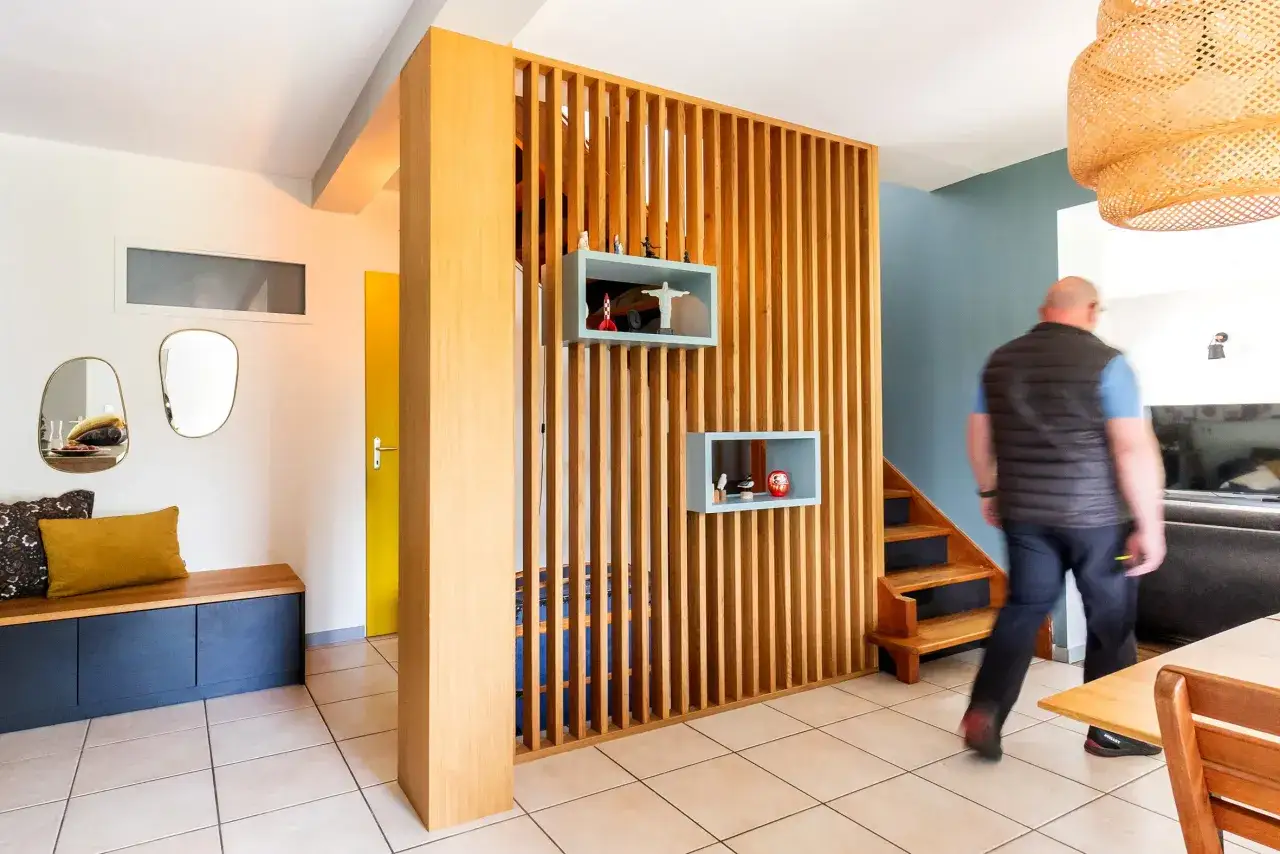 Un homme traverse un espace de vie moderne avec un s&eacute;parateur en bois, rappelant un atelier menuiserie maison. Des objets d&eacute;coratifs sont sur des &eacute;tag&egrave;res.