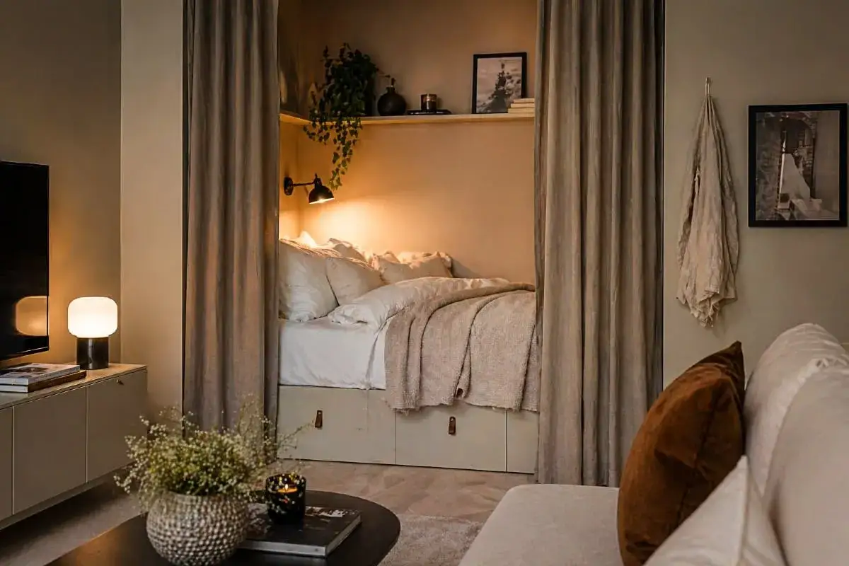 Mysig liten alkov med en inbjudande säng, draperad med mjuka textilier. En vägglampa ger ett varmt sken.