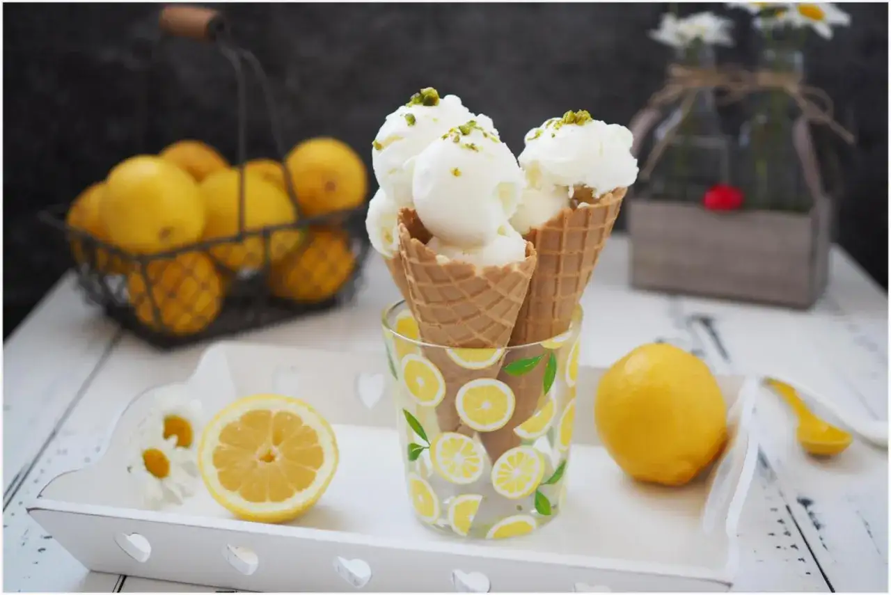 Erfrischendes Zitroneneis selber machen: Drei Waffeln mit cremigem Eis, garniert mit Pistazien, stehen in einem Glas mit Zitronenmuster. Frische Zitronen und Blumen im Hintergrund.