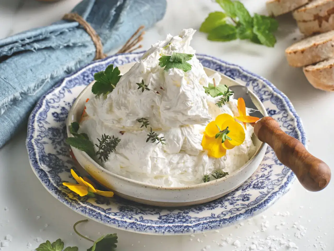 Burro montato soffice e arioso, guarnito con erbe aromatiche e fiori gialli, pronto per essere spalmato su pane croccante.