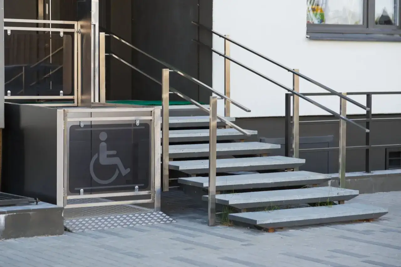 Piattaforma elevatrice senza cabina con simbolo disabili, accanto a una scala esterna con corrimano in metallo.
