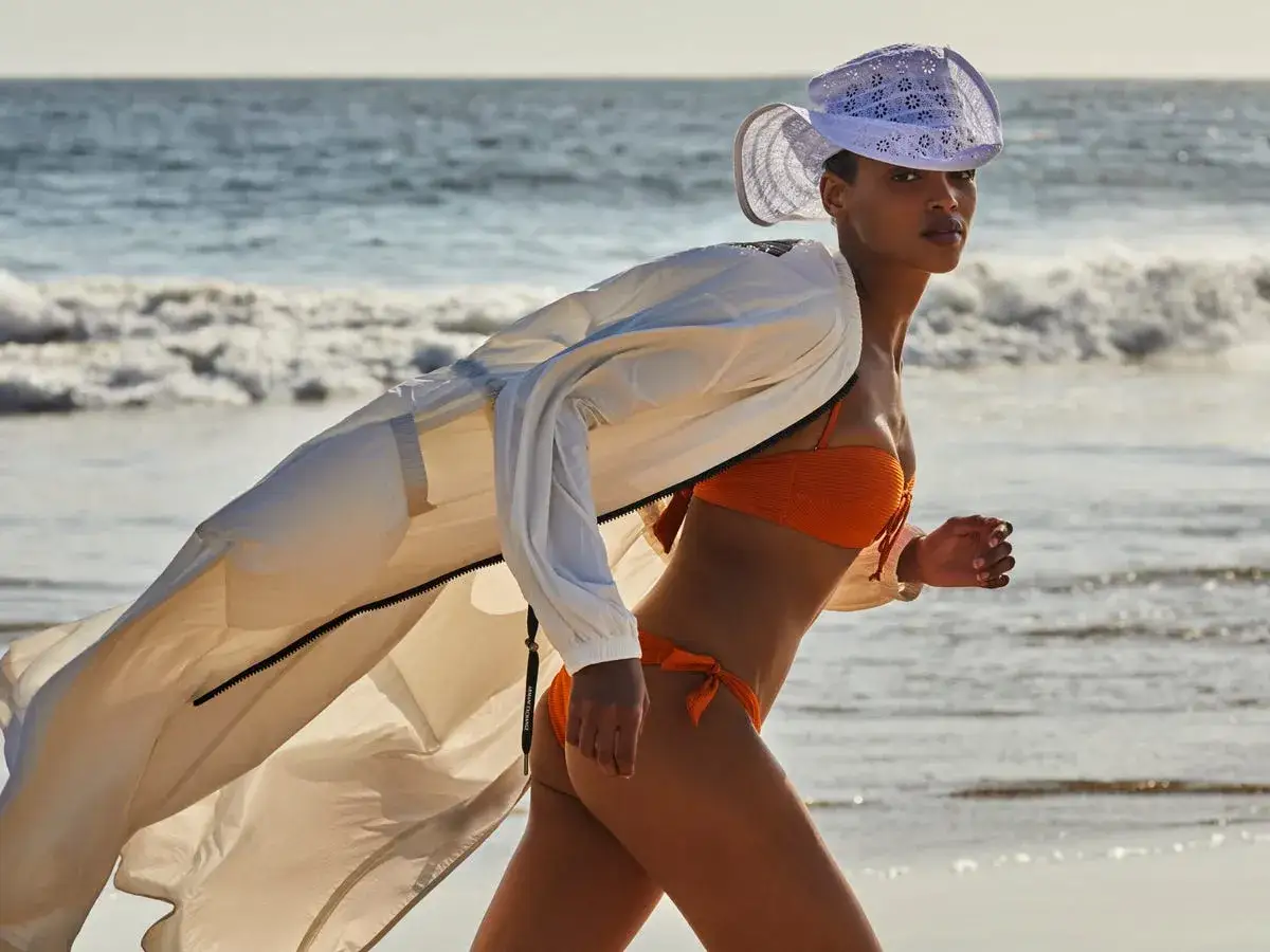 Donna in costume arancione corre sulla spiaggia, con un leggero giubbotto bianco mosso dal vento.