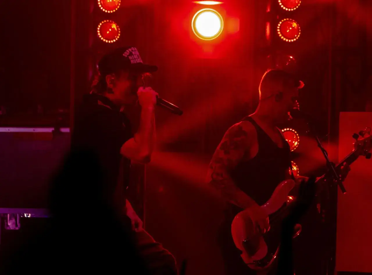 Un cantante con cappellino e un bassista tatuato sul palco, evocano l'energia dei gruppi punk anni 2000. Luci rosse intense.