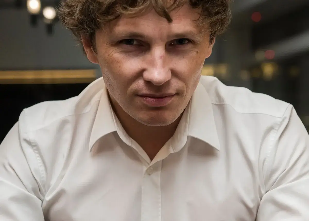 Mężczyzna w białej koszuli, z burzą blond włosów, patrzy wprost. Jego spojrzenie sugeruje, że za nim kryje się zaorski wiek doświadczeń.