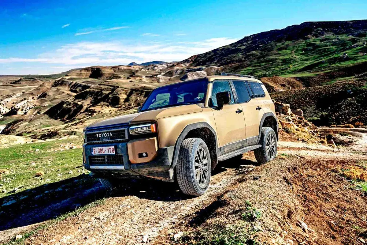 Nowy Toyota Land Cruiser w piaskowym kolorze pokonuje wyboisty teren na tle malowniczych, skalistych wzgórz.