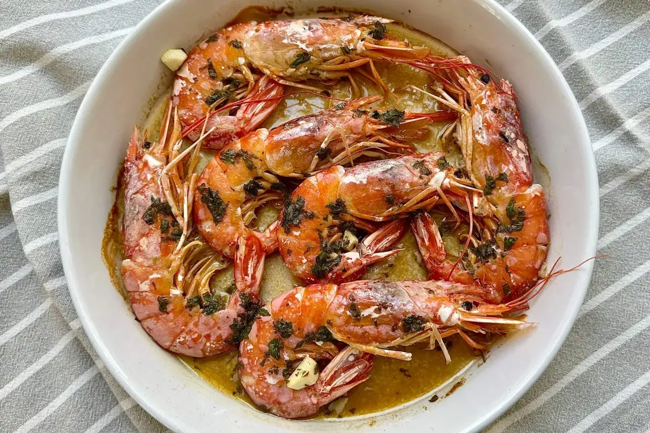 Gamberoni al forno succulenti, dorati e profumati con aglio e prezzemolo, serviti in una teglia bianca su un tovagliolo a righe.