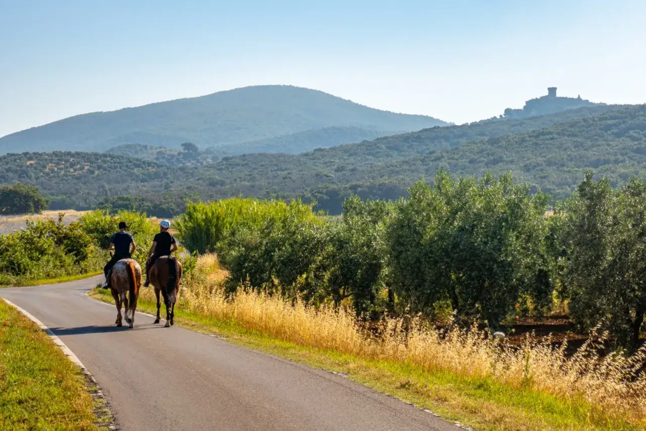 Due cavalieri percorrono una strada in un paesaggio collinare della Toscana, con ulivi e un castello in lontananza.
