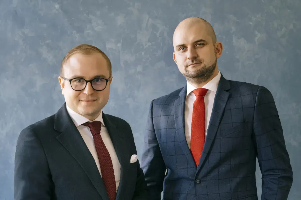 Dwóch mężczyzn w garniturach, jeden w okularach, na niebieskim tle.