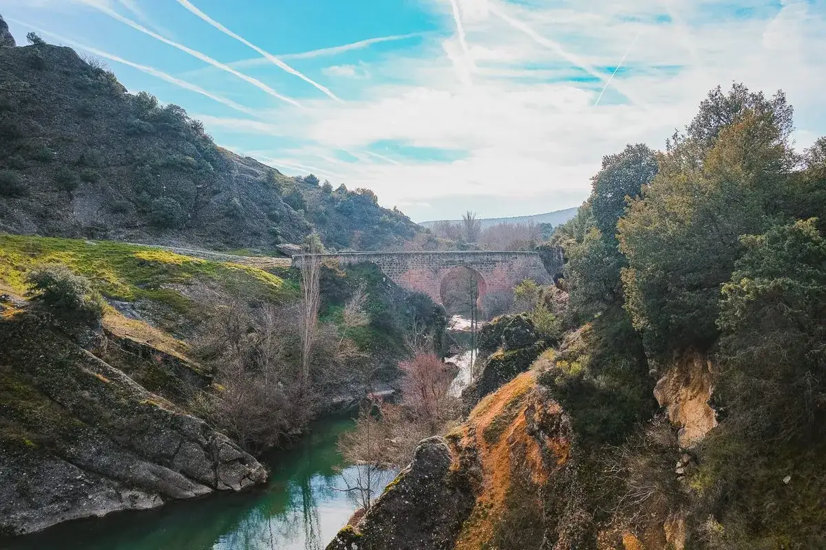Un puente de piedra cruza un r&iacute;o verde en un ca&ntilde;&oacute;n rocoso. El cielo azul tiene estelas blancas. Se dice que este lugar es la poza de do&ntilde;a urraca.