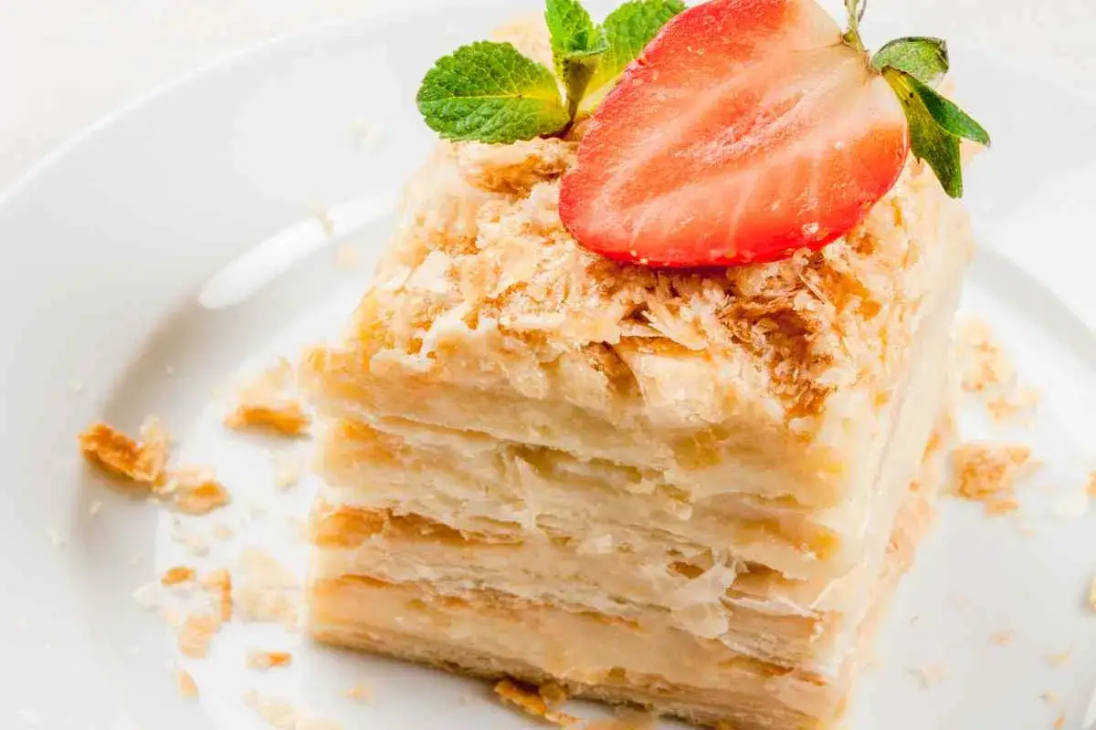 Fetta di torta millefoglie con strati di pasta sfoglia e crema pasticcera con panna, guarnita con fragola e menta.