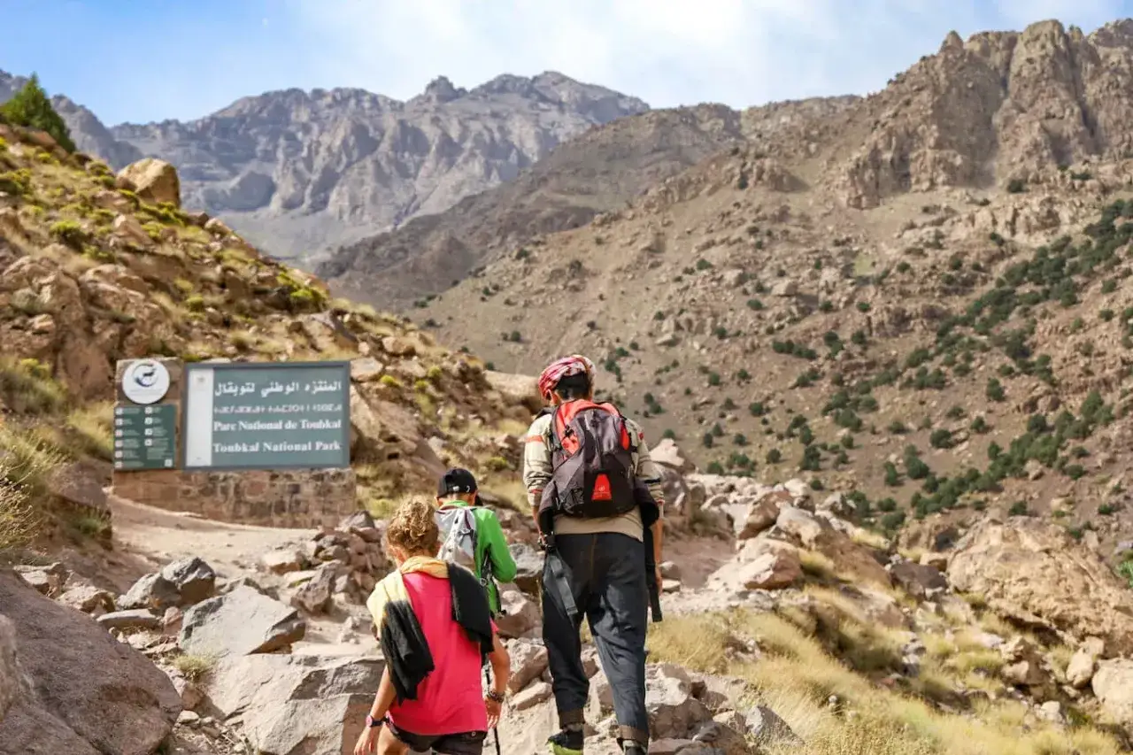 Turyści wędrują szlakiem w g&oacute;rach Atlas w Maroku, mijając tablicę Parku Narodowego Toubkal.