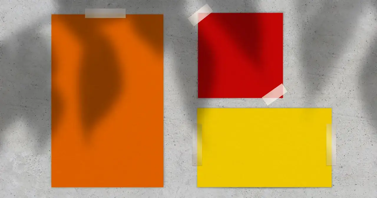 Drei bunte Papierbögen – orange, rot und gelb – sind auf einer Betonwand befestigt. Sie scheinen sich zu vermischen und ein warmes Farbspiel zu erzeugen.