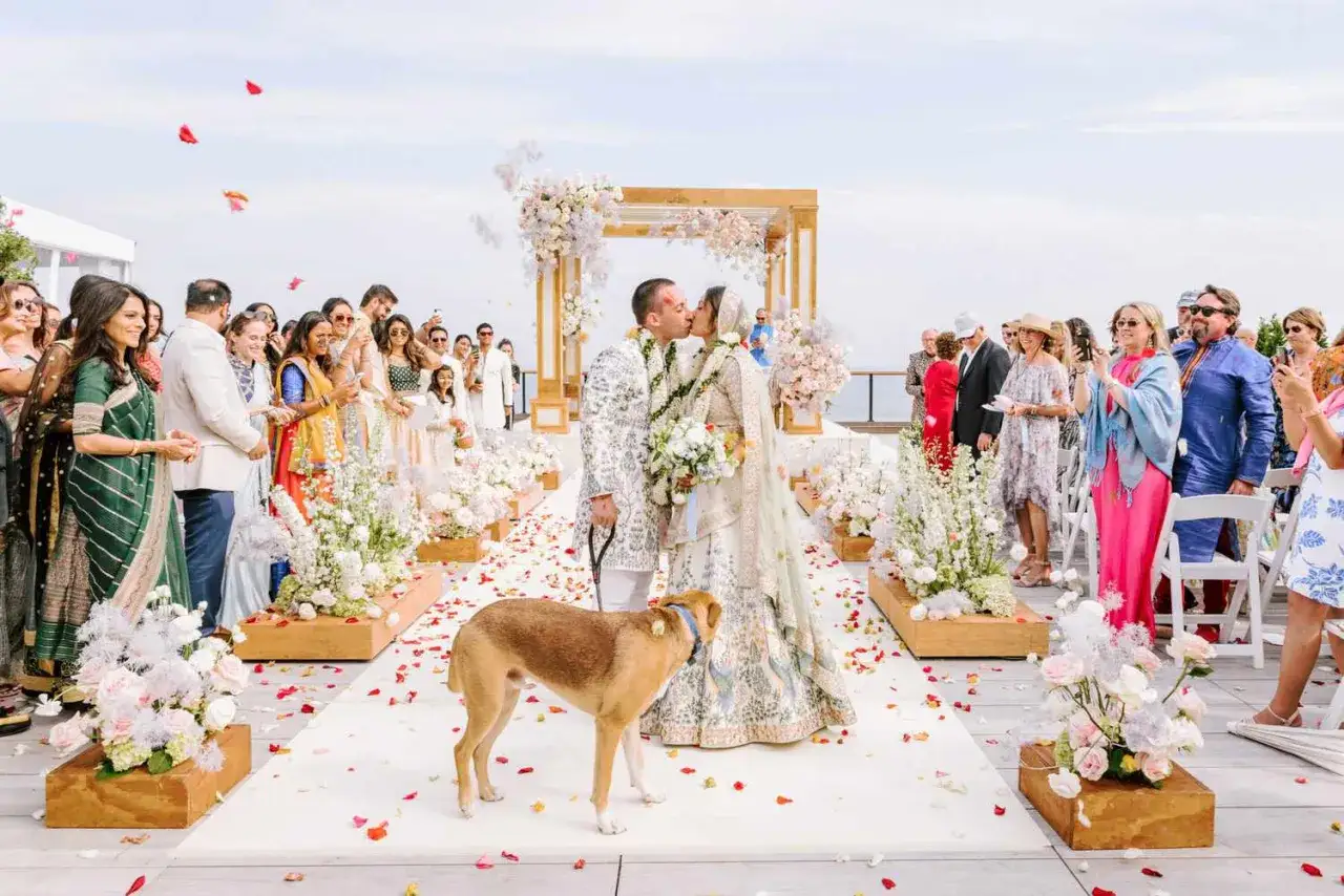 Una pareja se besa en su boda simb&oacute;lica al aire libre, rodeada de invitados y flores. Un perro observa la escena.