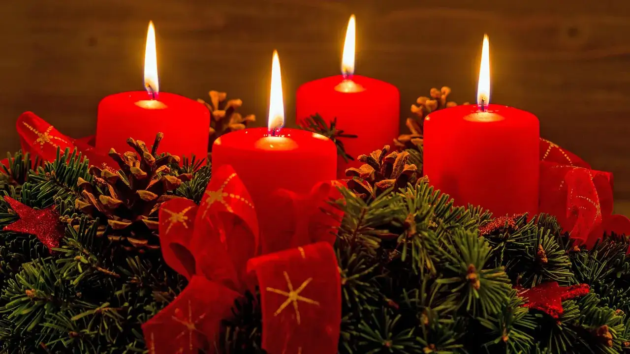 Vier brennende rote Kerzen auf einem Adventskranz mit Tannenzweigen, Zapfen und roten Bändern.