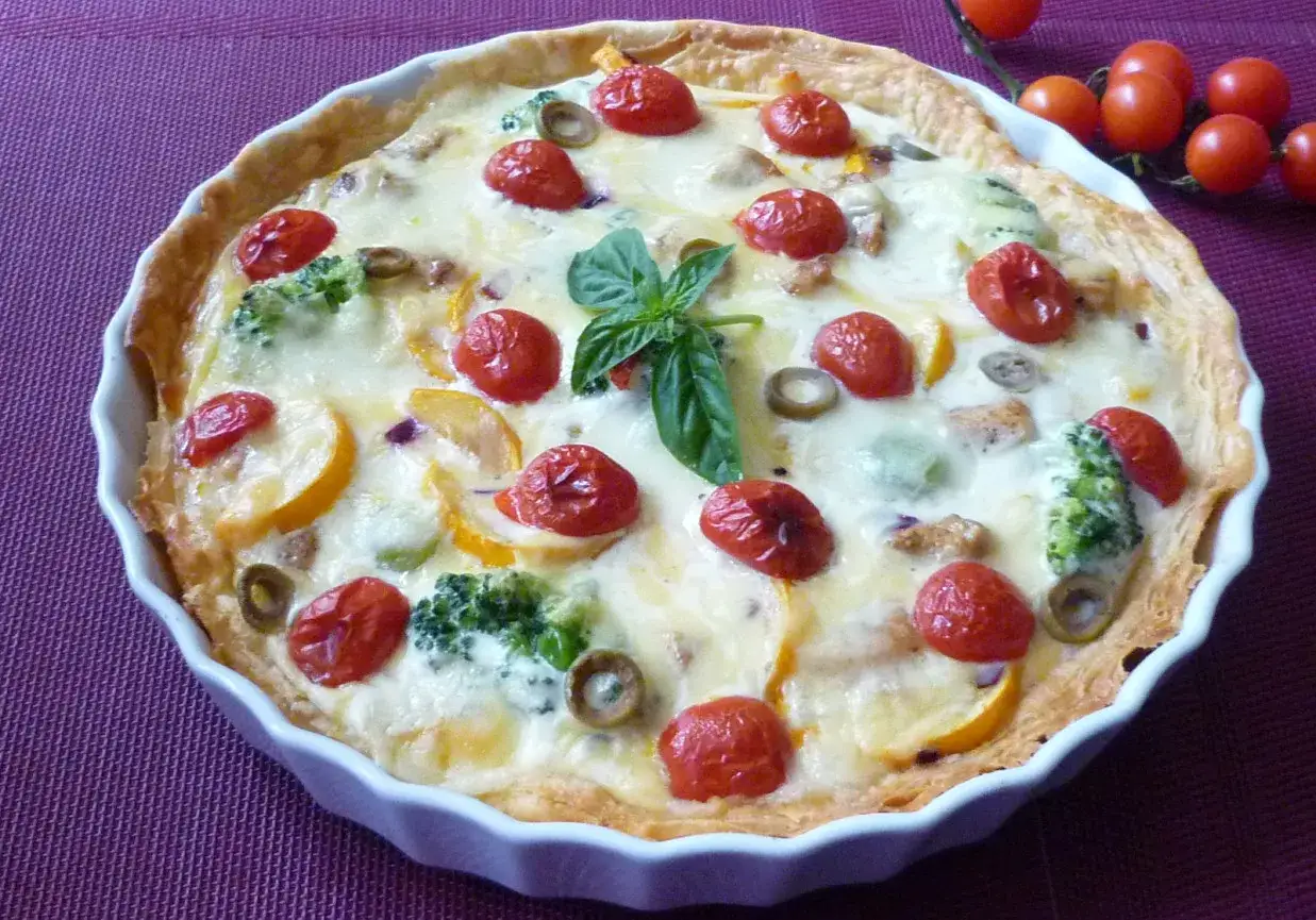 Pyszna tarta z kurczakiem, brokułami, pomidorkami i oliwkami, udekorowana listkiem bazylii.