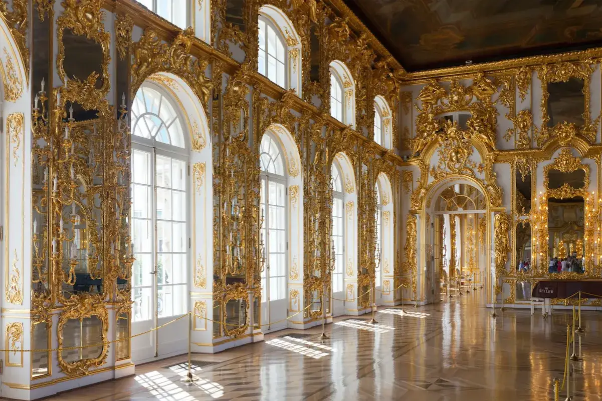 An opulent hall adorned with intricate gold stucco work in the Rococo style, featuring tall arched windows and a polished wooden floor reflecting the sunlight.