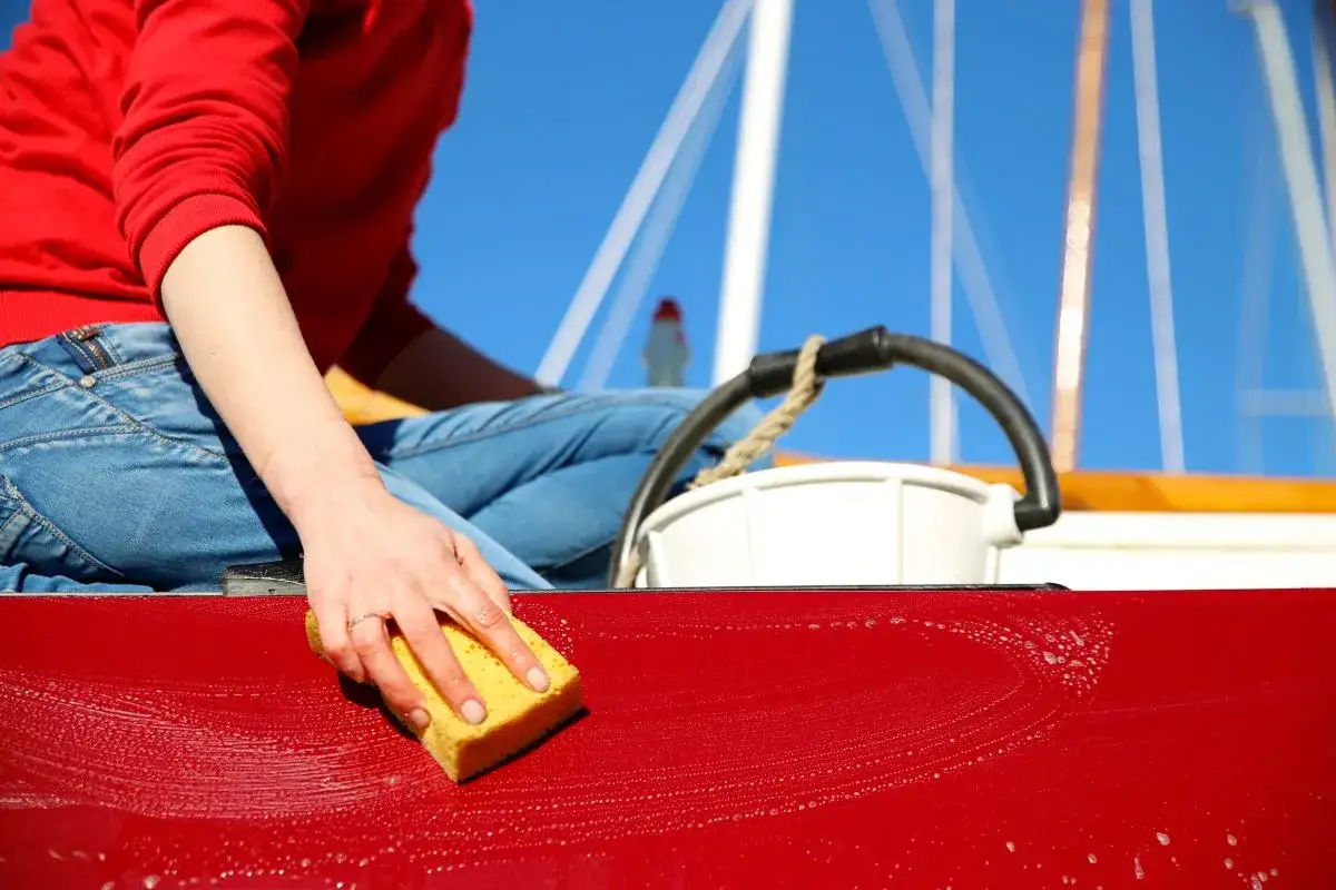 Main d'une personne faisant la mise en cire d'une surface rouge avec une éponge, sur un bateau par temps ensoleillé.