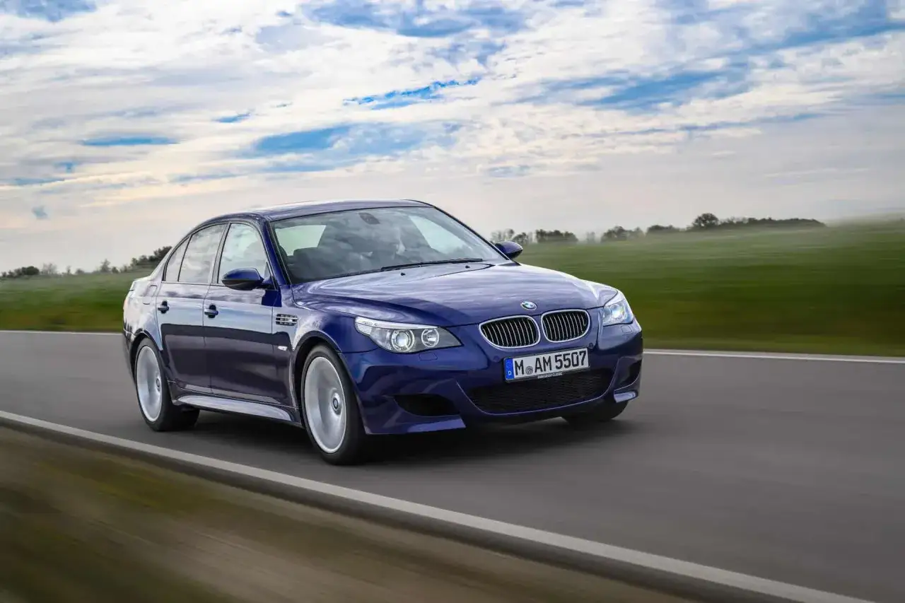 Un BMW M5 E60 azul oscuro acelera por una carretera rural, con campos verdes y un cielo nublado de fondo.