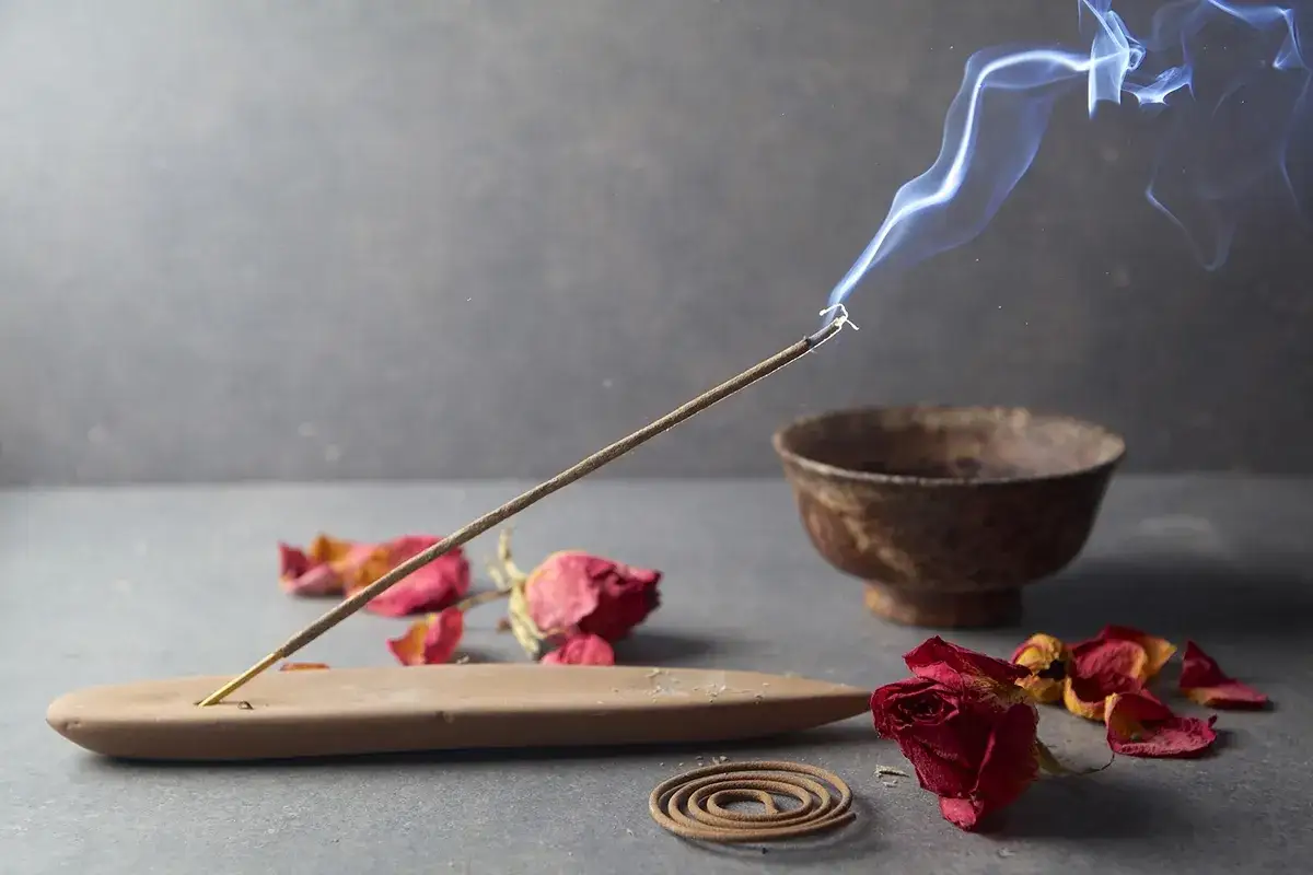 Humo azul se eleva de un incienso purificador, rodeado de pétalos de rosa secos y un cuenco rústico.
