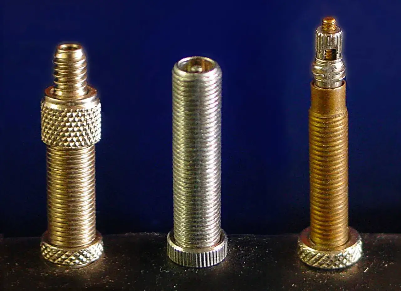 Drei Ventile für Fahrradschläuche, nebeneinander auf dunklem Grund. Ein goldfarbener, ein silberner und ein gold-silberner Fahrradschlauch automat.