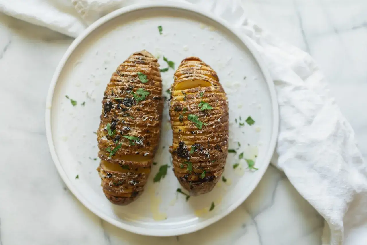 Deux pommes de terre hasselback dorées, coupées en fines lamelles et garnies de persil frais et de fromage râpé, sur une assiette blanche.