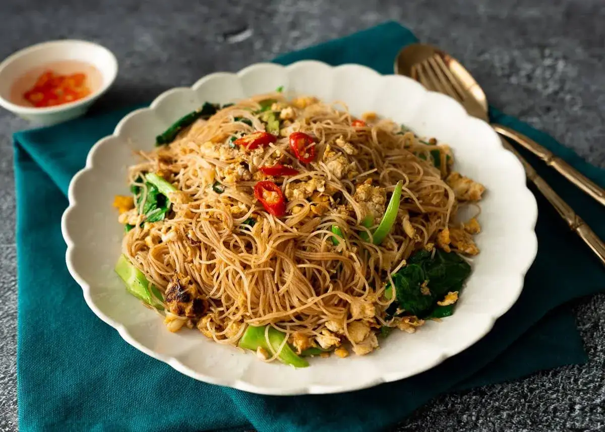 Un piatto invitante di spaghetti di riso saltati con verdure, uova e peperoncino, servito su un piatto bianco con un contorno di salsa.
