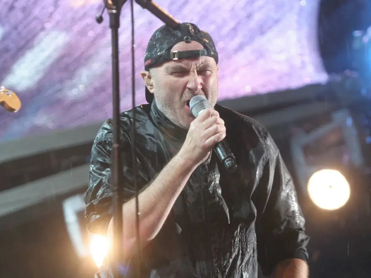 Phil Collins, w czapce i mokrej kurtce, śpiewa do mikrofonu na koncercie. To legendarny wokalista zespołu Genesis.
