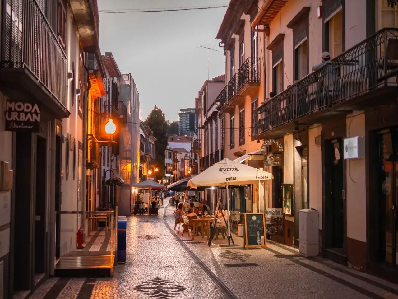 Ulica w Funchal, stolicy Madery, wieczorem. Widać restauracje z ogródkami, sklepy i ludzi spacerujących po brukowanej ulicy.