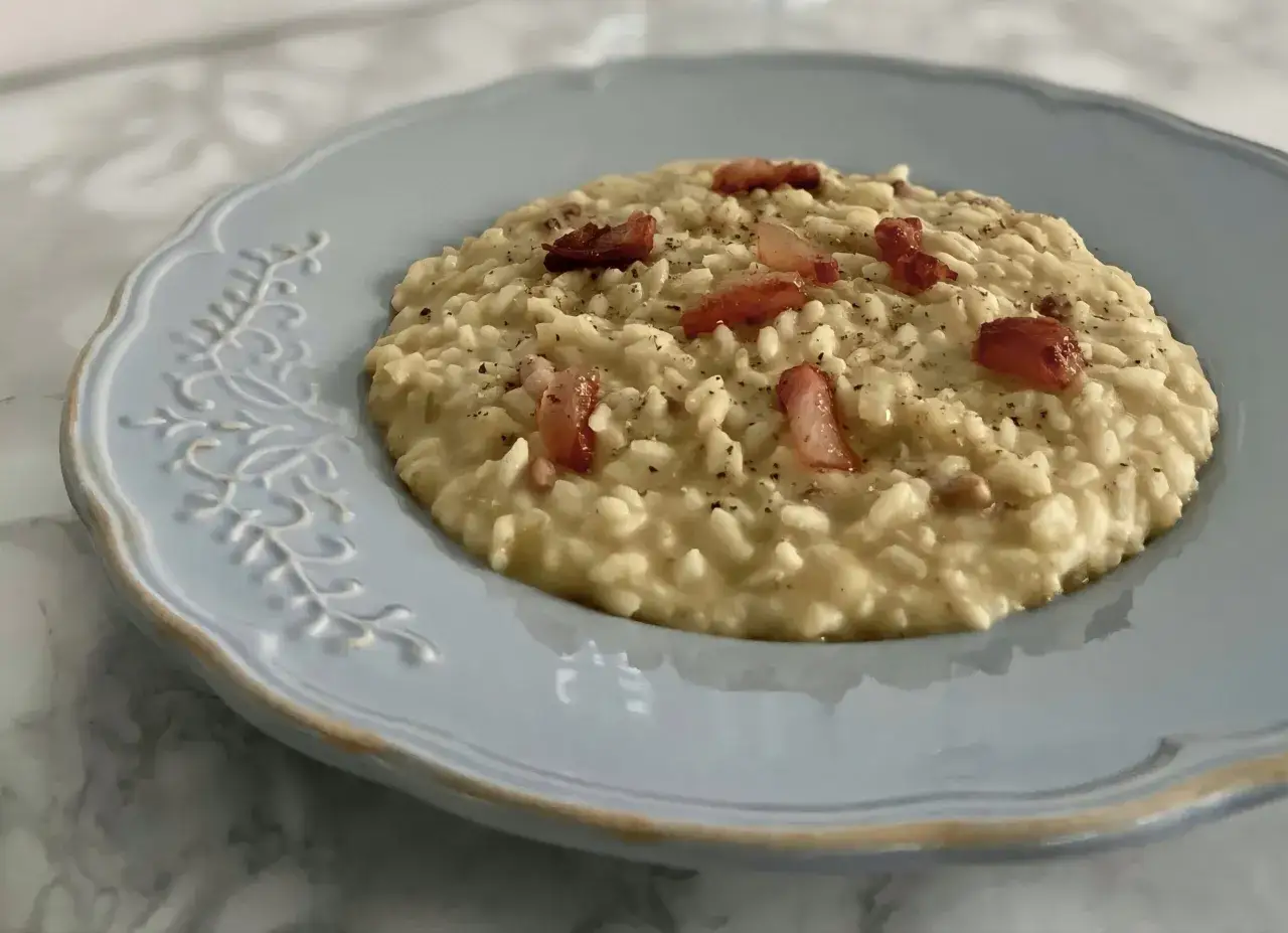 Un cremoso risotto con ceci e pancetta croccante, servito su un piatto azzurro decorato.