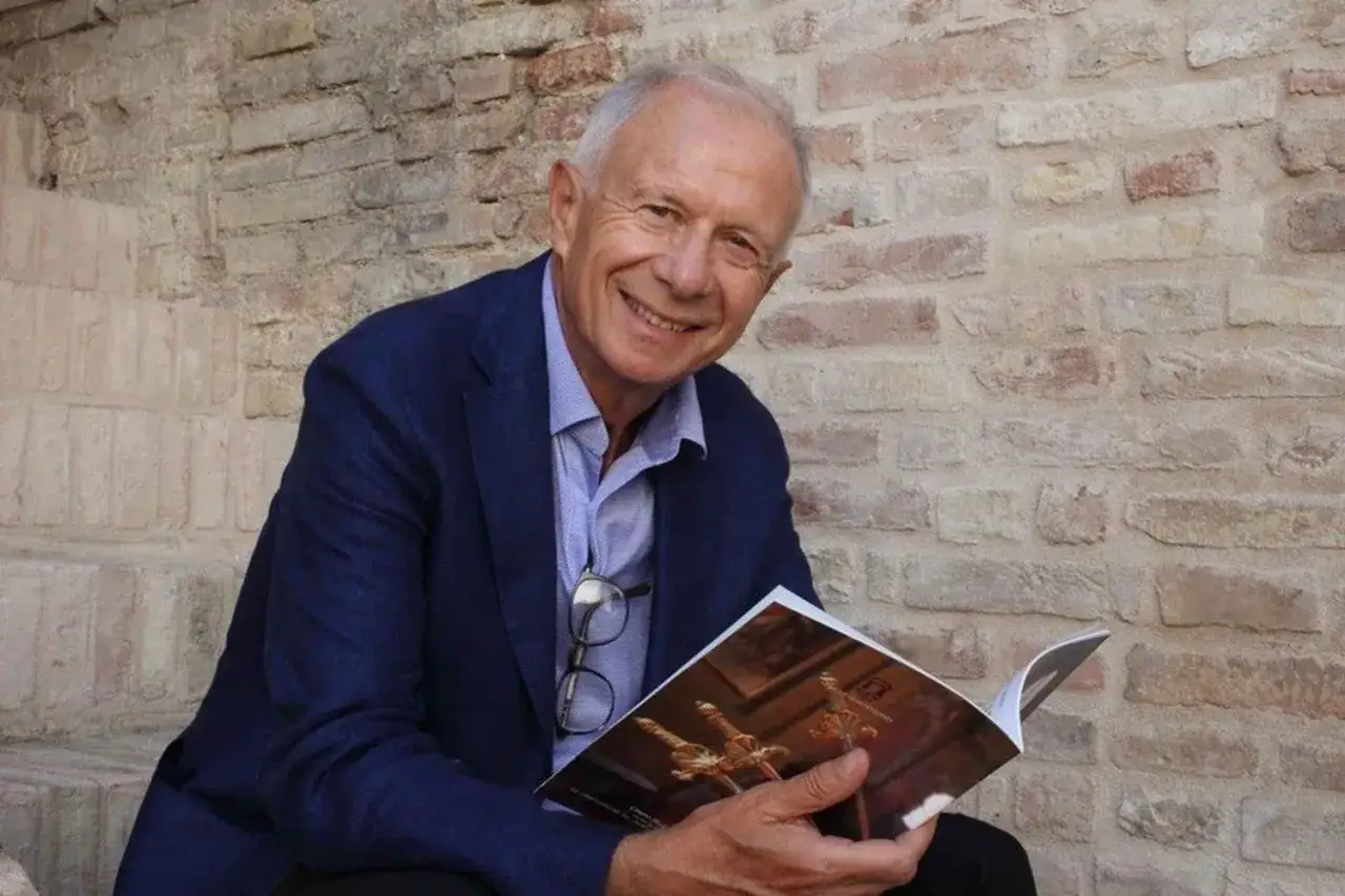 Padre Alberto Maggi sorride con un libro aperto, appoggiato a un muro di mattoni.