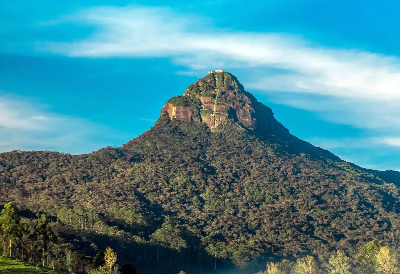 Adams Peak reser sig majest&auml;tiskt, kl&auml;dd i gr&ouml;nska, med en helgedom p&aring; toppen under en klarbl&aring; himmel.