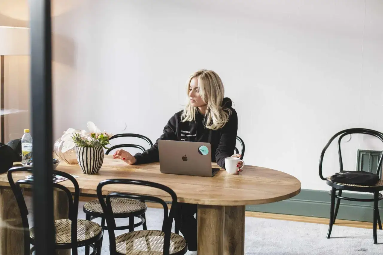 En kvinna med blont h&aring;r sitter vid ett tr&auml;bord med en laptop och en kaffekopp. Hon verkar fundera, kanske p&aring; grund av samarbetssv&aring;righeter med en kollega.