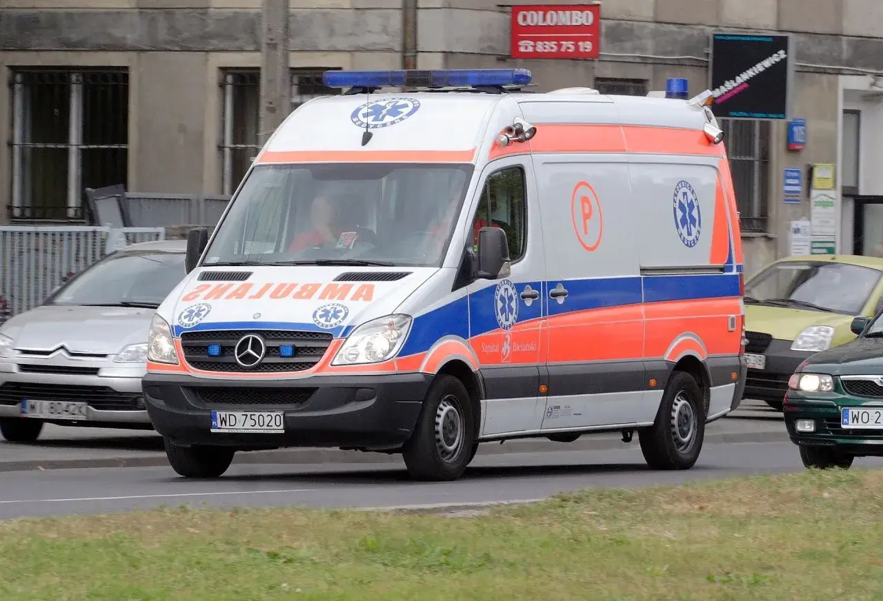 Biała karetka z czerwono-niebieskimi pasami i oznaczeniami medycznymi jedzie ulicą. Widoczne są numery rejestracyjne i nazwa szpitala.