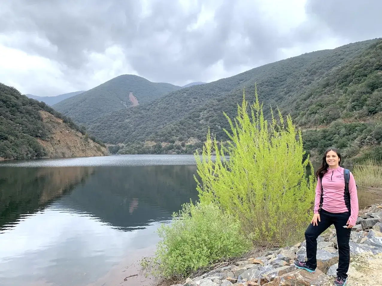 Mujer sonr&iacute;e junto a un lago sereno, rodeado de monta&ntilde;as verdes. El paisaje evoca la tranquilidad de la panta de Vallforners.