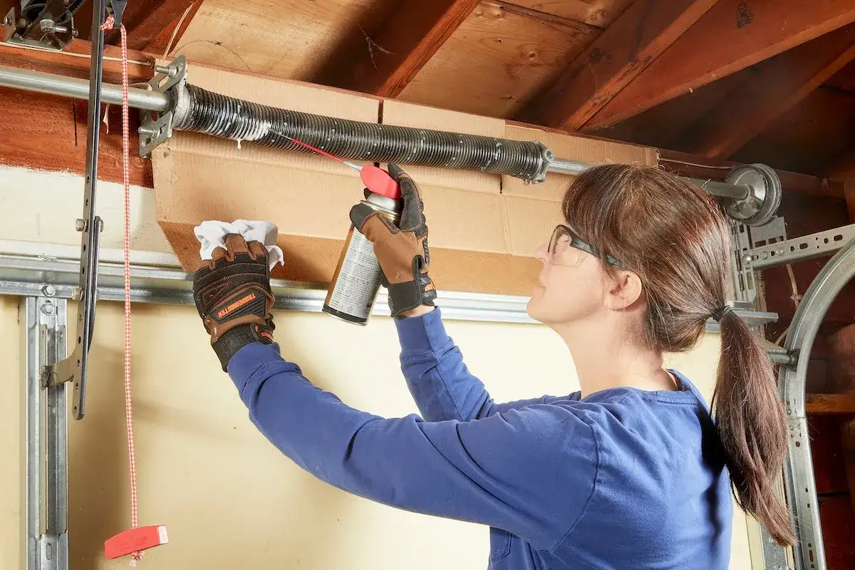 A person in gloves sprays garage door lube onto a spring while holding a rag.