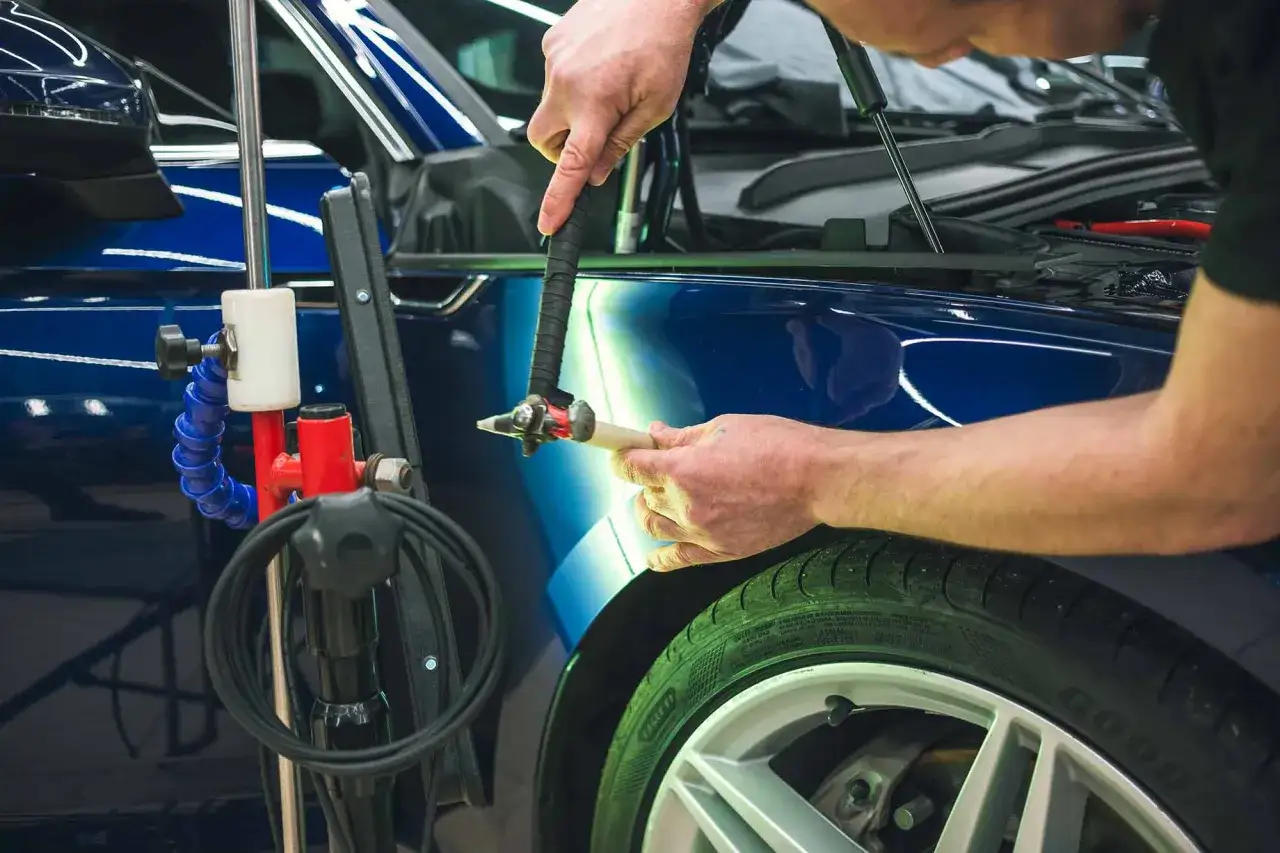 A technician uses a specialized tool to repair hail damage on a blue car's fender.