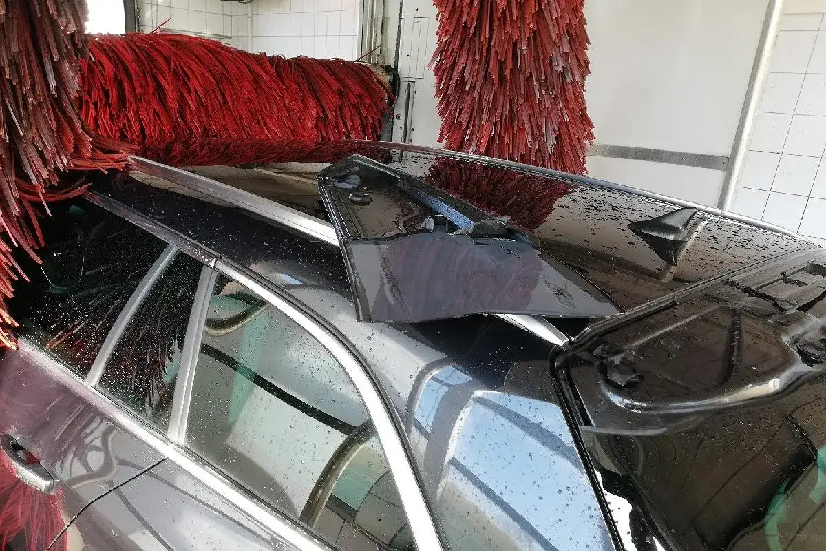 Czarne auto w myjni automatycznej, czerwone szczotki wirują. Czy myjnia automatyczna niszczy lakier? Woda spływa po karoserii.