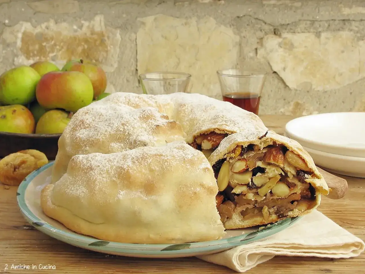 Strudel di mele e mandorle, una rocciata di Assisi, cosparso di zucchero a velo, con mele fresche sullo sfondo.