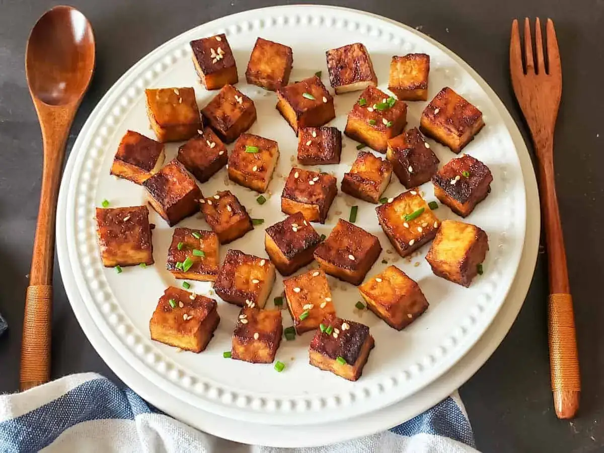 Chrupiące kostki tofu, idealne do curry, posypane sezamem i szczypiorkiem, podane na białym talerzu z drewnianymi sztućcami.