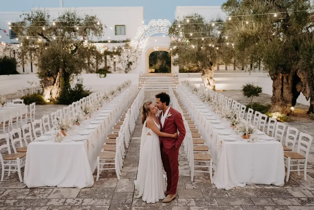 Un couple s'embrasse devant de longues tables dress&eacute;es pour un mariage dans les Pouilles, sous des guirlandes lumineuses et des oliviers centenaires.