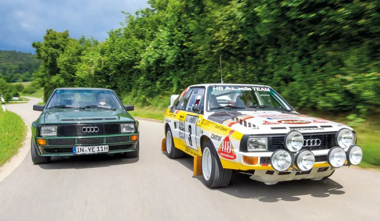 Dwa kultowe Audi Sport Quattro pędzą po krętej drodze. Jeden zielony, drugi w barwach rajdowych.