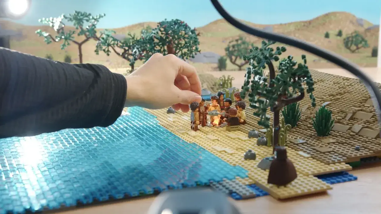 Eine Hand platziert Figuren für ein **Lego Stop Motion** am Strand. Eine Gruppe von Minifiguren versammelt sich um ein Lagerfeuer.