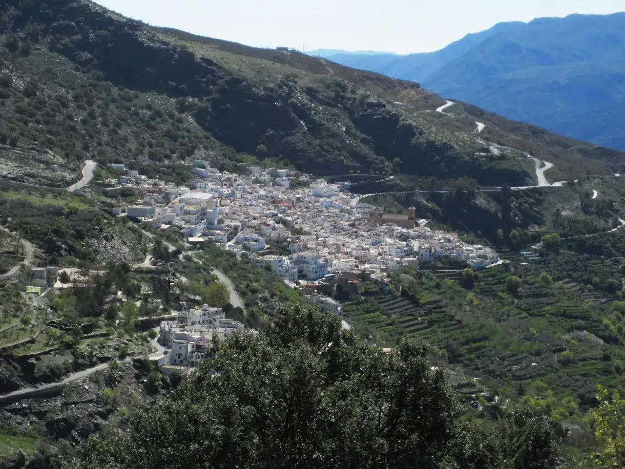 Pueblo blanco en ladera montañosa, con caminos sinuosos que recuerdan a un **río chico** serpenteando.