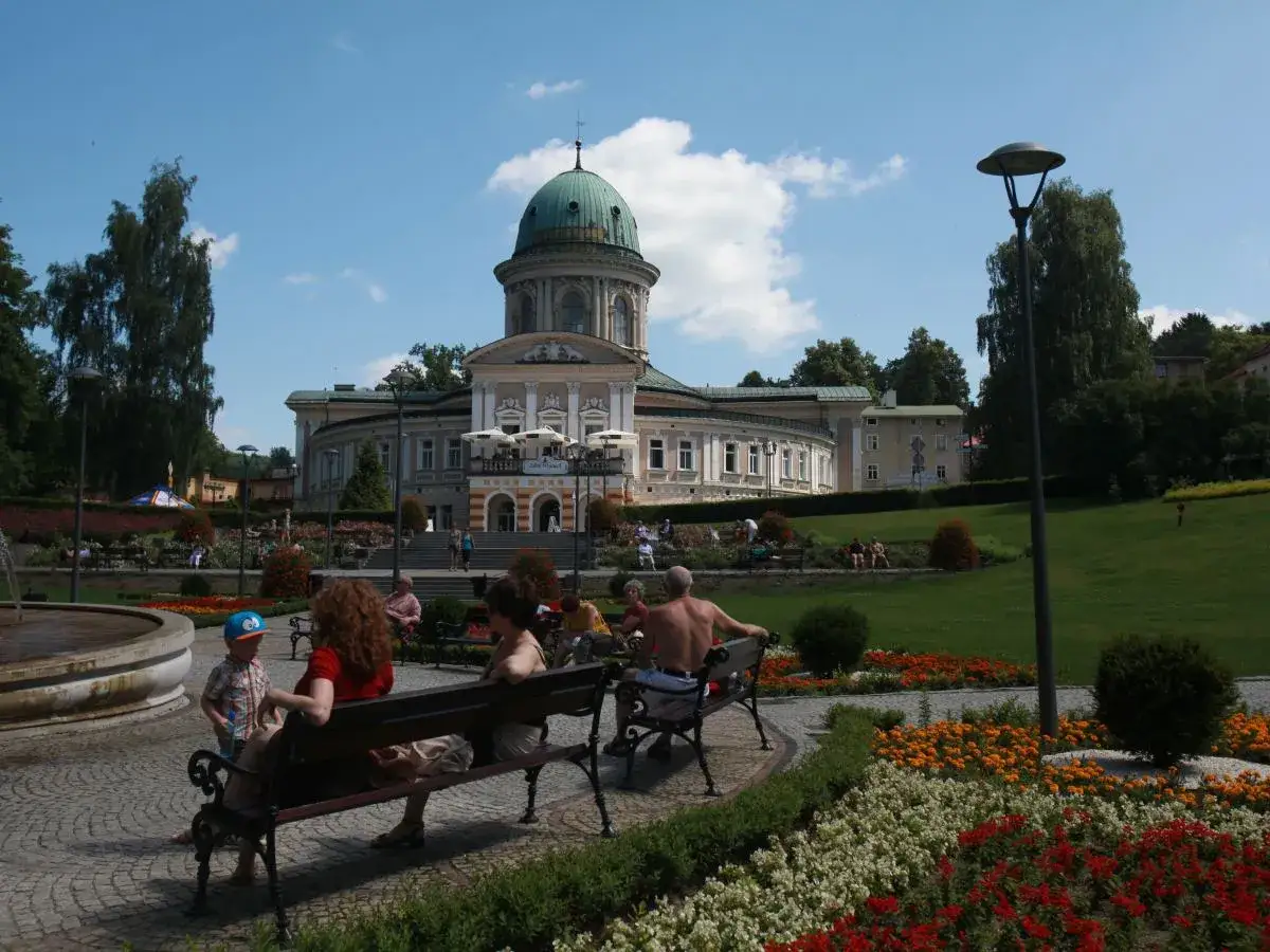 Uzdrowisko w Kotlinie Kłodzkiej. Ludzie odpoczywają na ławkach w pięknym parku z fontanną i kwitnącymi rabatami kwiatowymi, w tle zabytkowy budynek z kopułą.