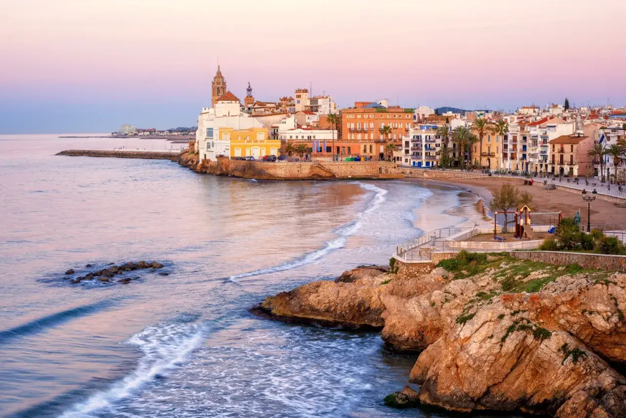 Zach&oacute;d słońca nad malowniczą plażą w Sitges, niedaleko Barcelony. Kolorowe budynki, morze i skaliste wybrzeże tworzą idylliczny krajobraz.