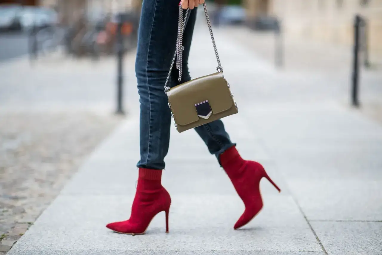 Une femme porte un jean slim et des bottines rouges &agrave; talons aiguilles, tenant un sac &agrave; main kaki.
