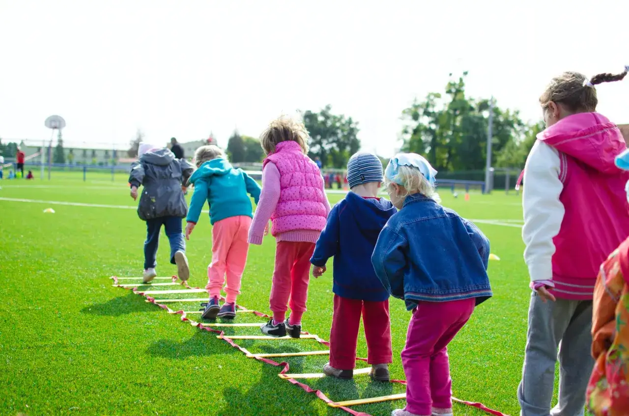 Ni&ntilde;os participan en actividades infantiles, corriendo en una escalera de agilidad en un campo de c&eacute;sped artificial.