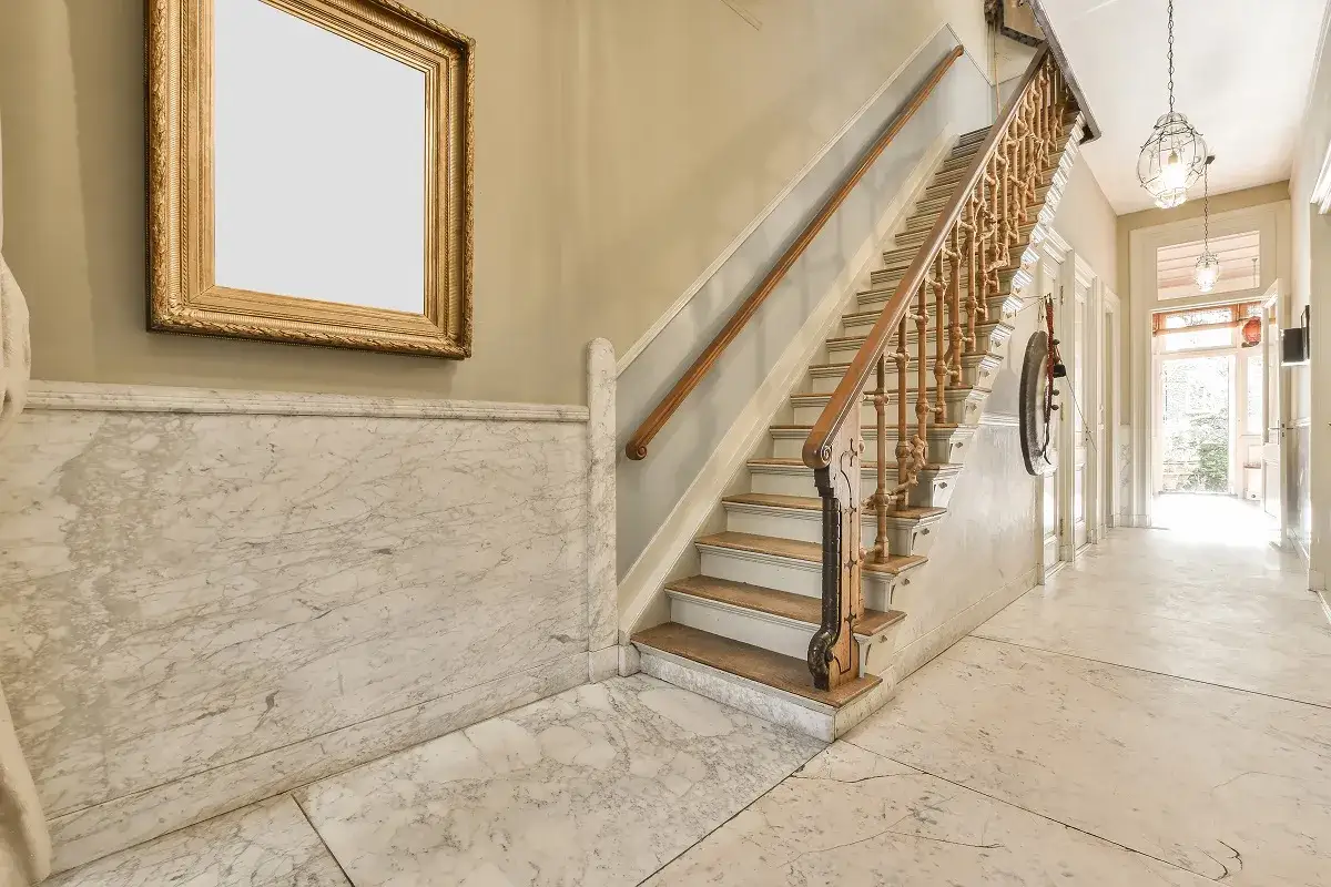 Amplio rellano de edificio con escalera de madera, m&aacute;rmol y un cuadro dorado.