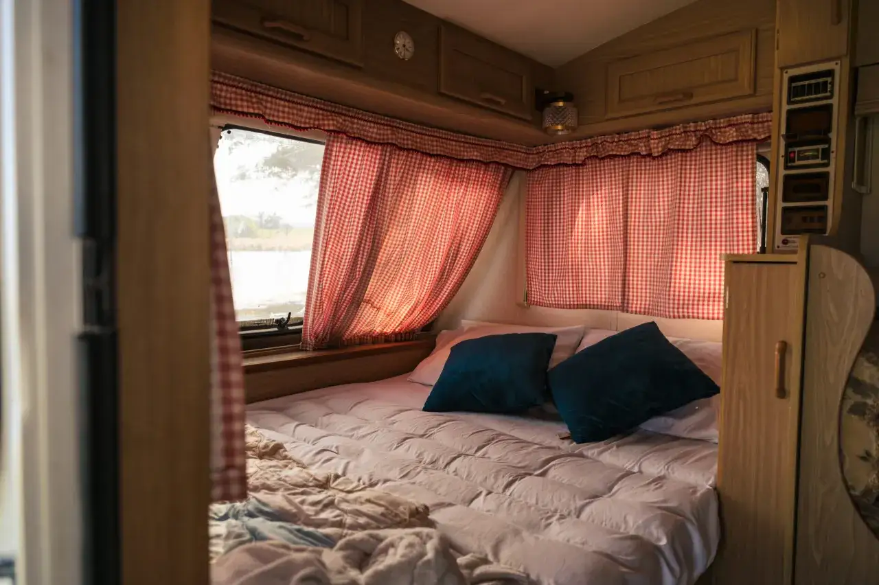 Intérieur d'un camping-car 7m avec lit central douillet, rideaux à carreaux rouges et coussins bleus. Prêt pour une nuit paisible.