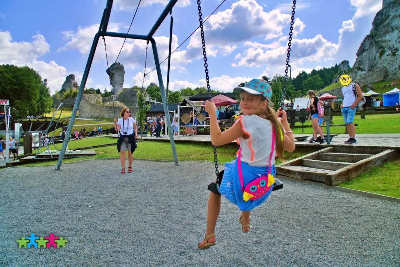 Dziewczynka na huśtawce w **Ogrodzieńcu park rozrywki**. W tle ruiny zamku i inni turyści.